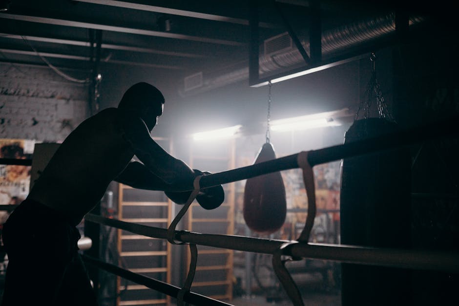 Moody silhouette of a boxer leaning in a dimly lit gym with punching bag and boxing ring